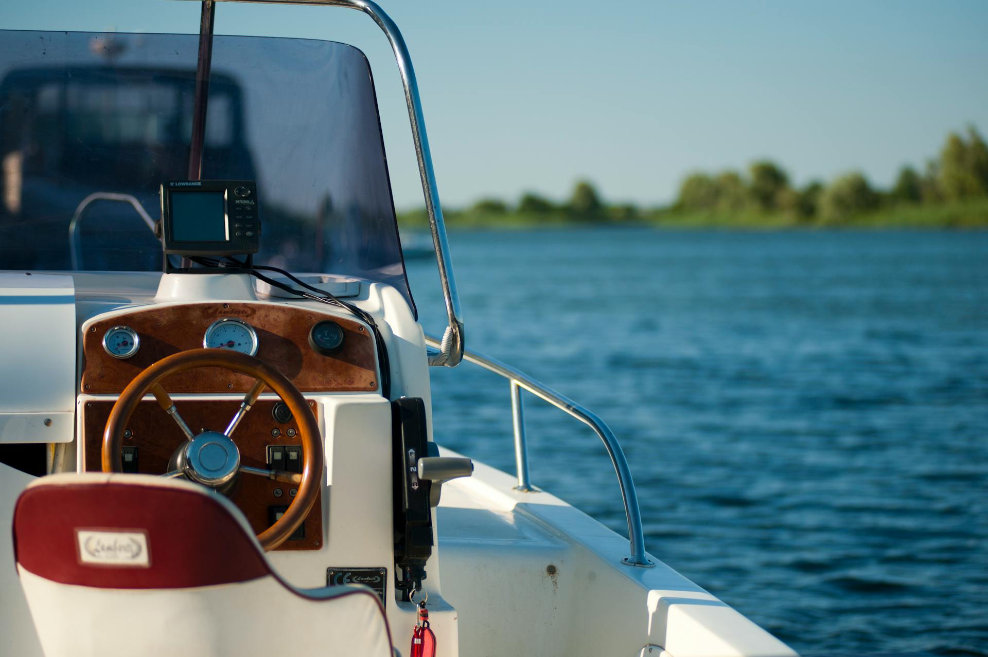 The helm of a boat with a wooden steering wheel, set against a calm body of water and green shoreline.